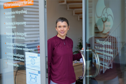 Frau steht freundlich an der Glastür eines Hörakustik-Fachgeschäfts mit Öffnungszeiten.